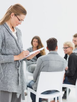 Serious female therapist writing notes with group therapy in session in background